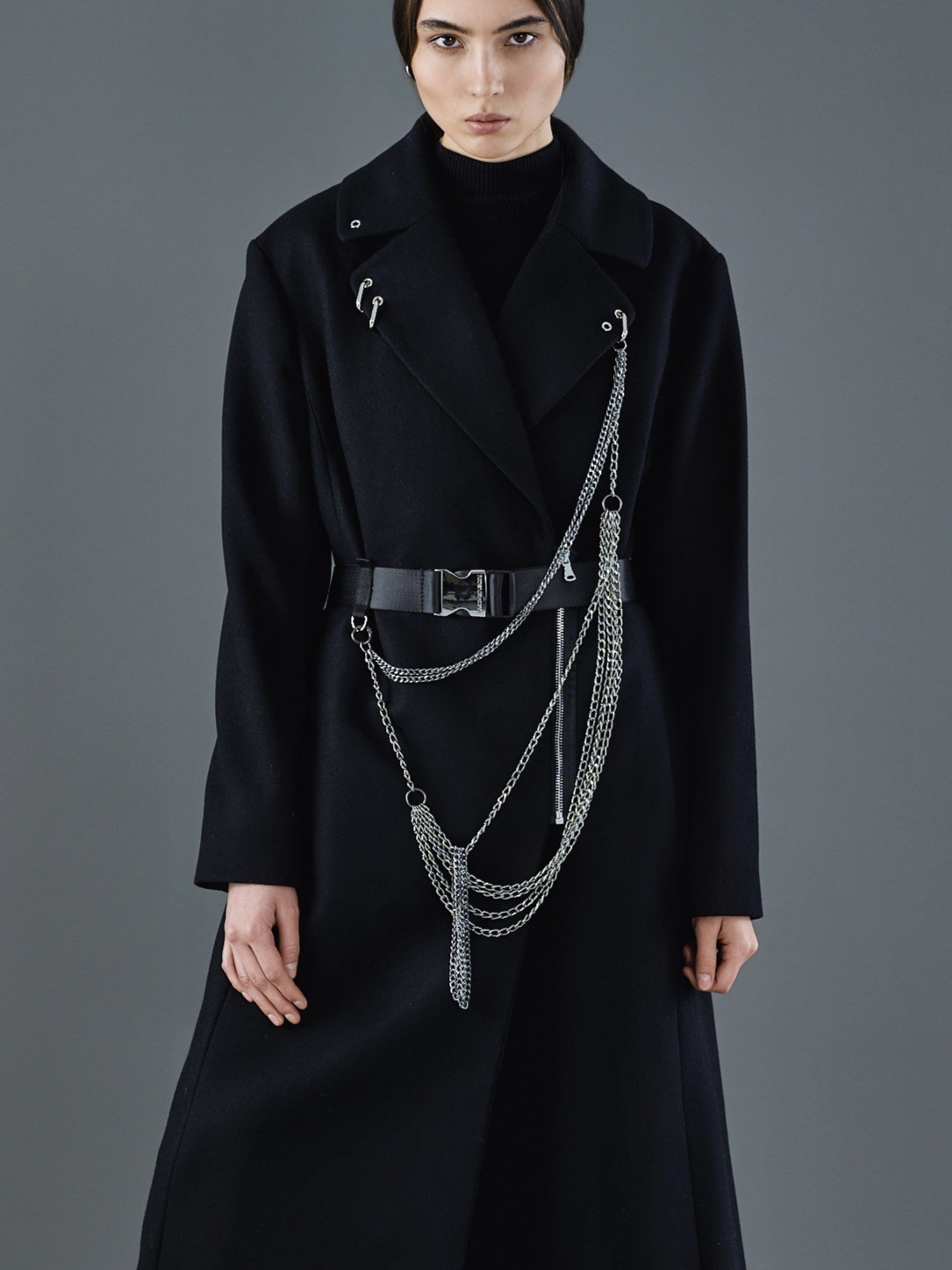 Женщина в черном длинном пальто с металлической цепочкой и ремнем, позирует на сером фоне. Стильный минималистичный образ с акцентом на аксессуары в виде серебряных цепей и металлического декора