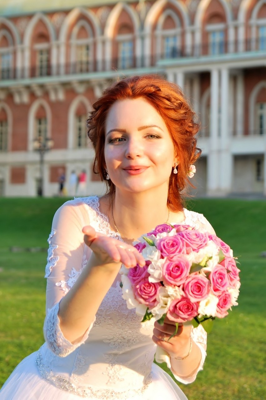 Свадебная фотосъёмка в Царицыно летом на закате, невеста посылает воздушный поцелуй на фоне дворца из красного и белого камня, в руках невеста держит бело-розовый букет