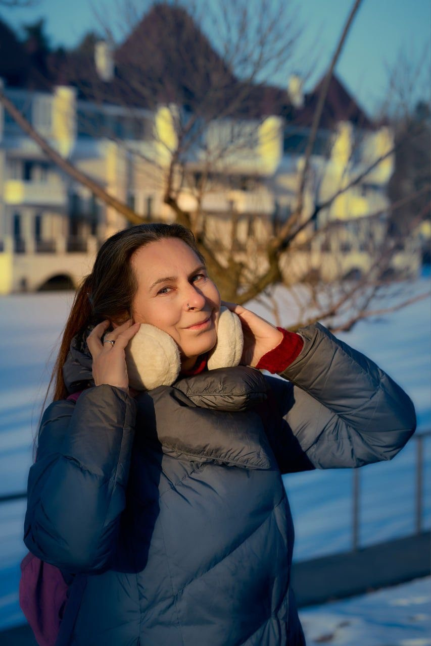Портрет улыбающейся женщины в темно-синей стеганой куртке и кремовых наушниках, стоящей на улице в снегу во время зимнего золотого часа.