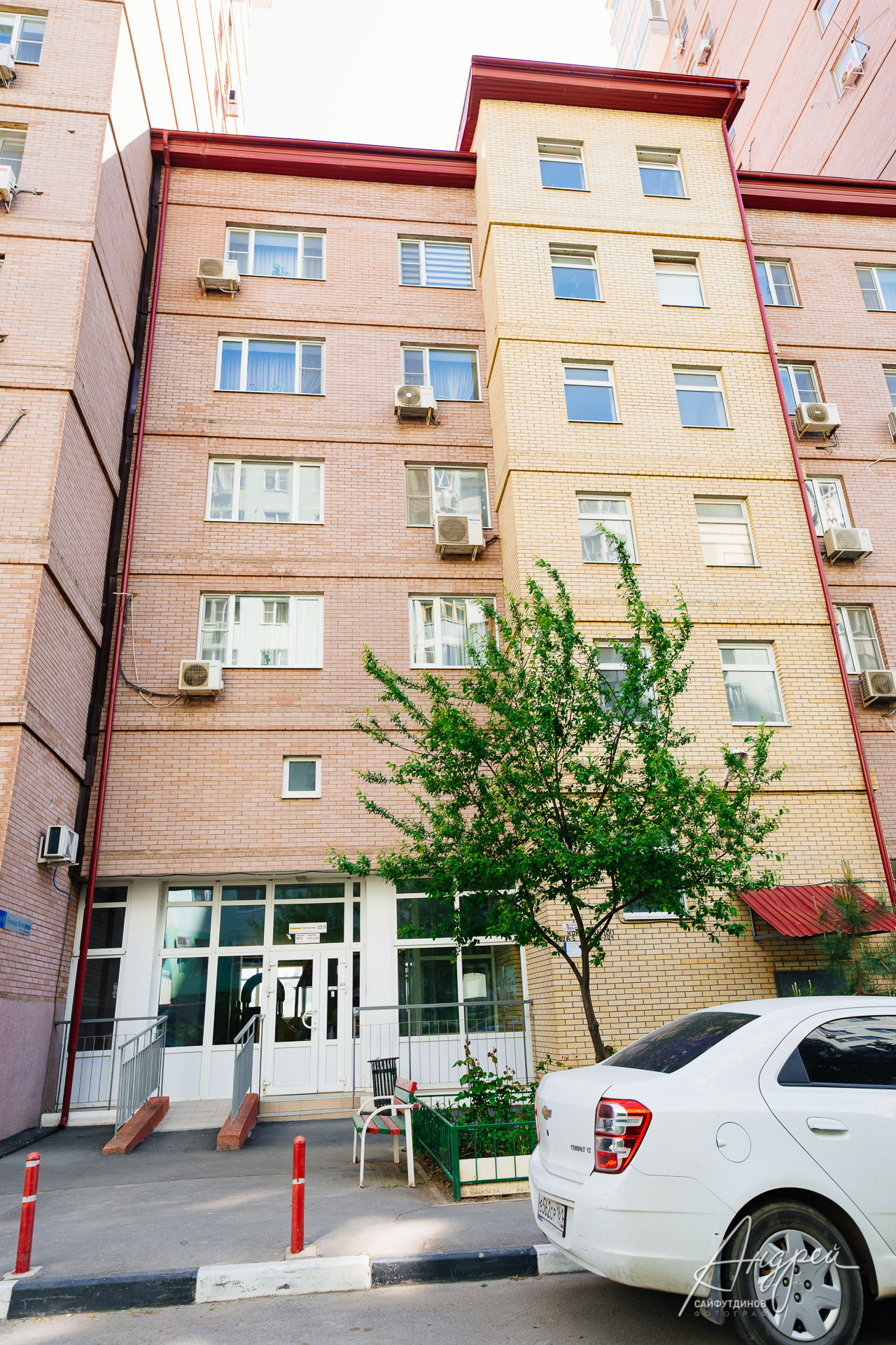 кирпичное здание с окнами и кондиционерами. Перед ним растёт дерево. Рядом стоит белый автомобиль.