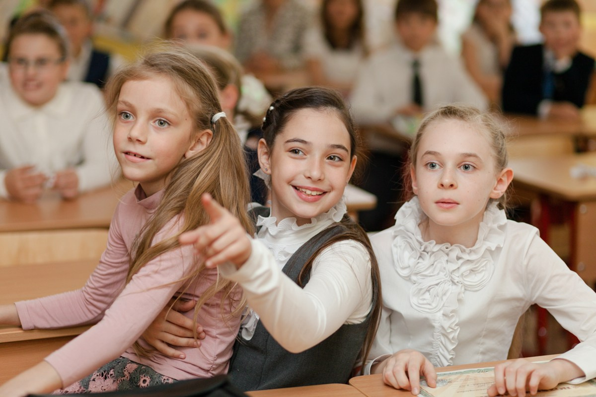 Три школьницы сидят за своей партой и улыбаются своему учителю