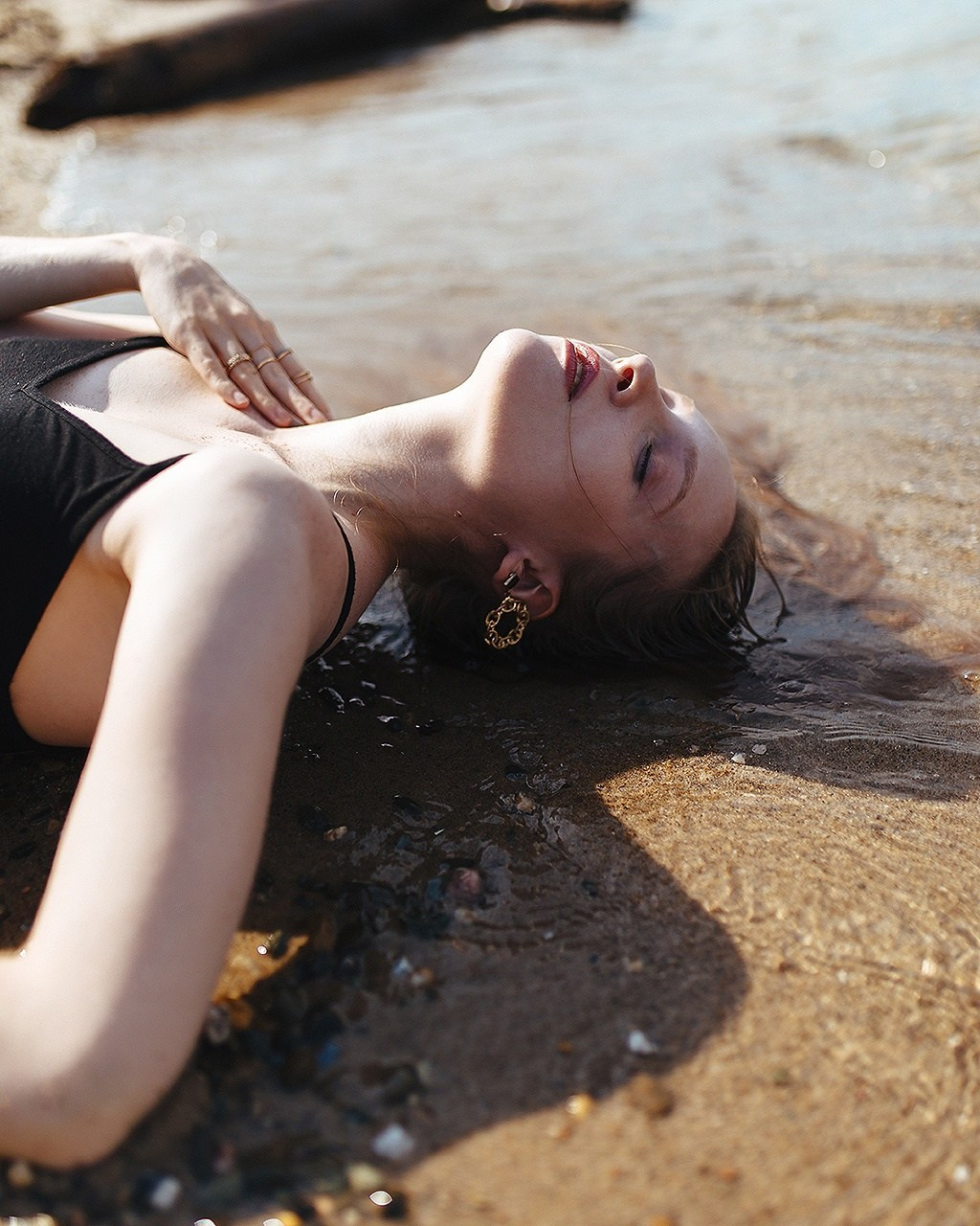 Портрет анфас девушки в черном платье и с крупными сережками в ушах, которая лежит на песке у воды, волосы в воде мокрые