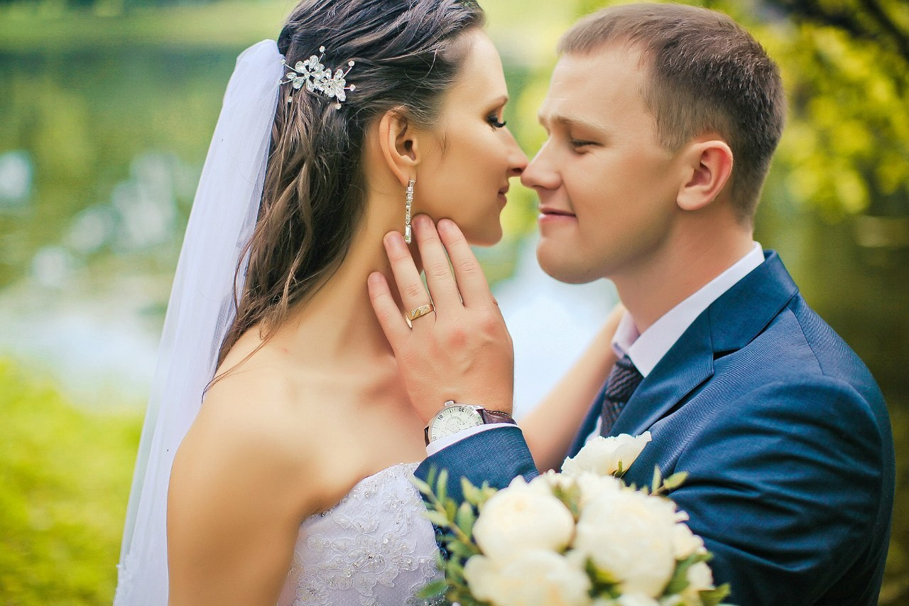 молодожены нежно касаются друг друга с белым букетом в руках на фоне пруда