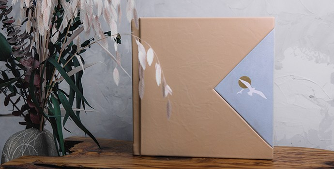 Квадратная фотокнига в бежевой кожаной обложке с голубой треугольной вставкой и тиснением чайки. Книга стоит на деревянном срезе в интерьере с сухоцветами.