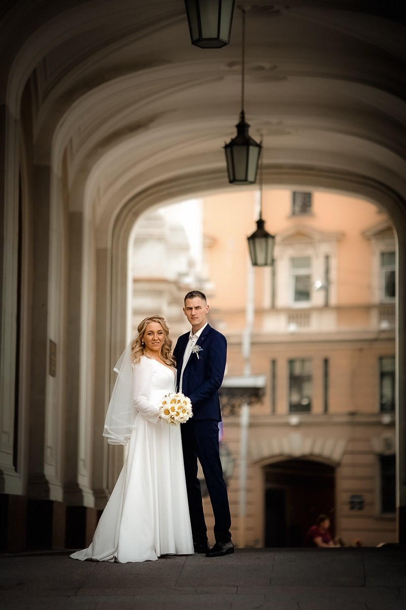 Жених и невеста смотрят в кадр, стоя на крыльце заксобрания в Санкт-Петербурге. Вертикальный ростовой кадр.