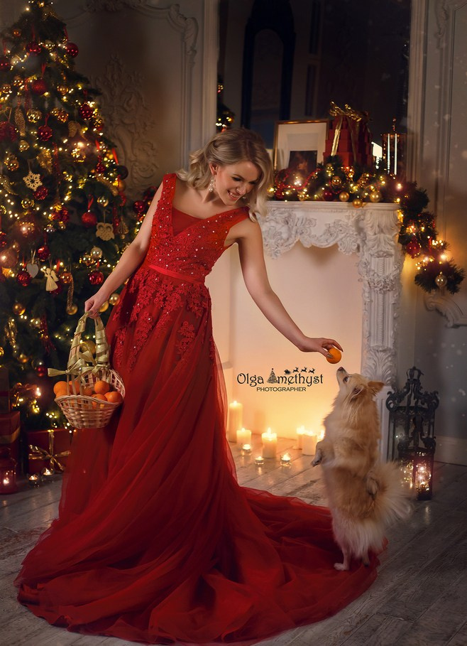 Красивая девушка в длинном красном платье дает мандарин собаке на новогодней фтосессии.