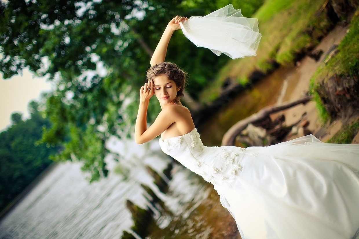 гибкая невеста в красивом белом платье держит в руке фату и позирует у реки