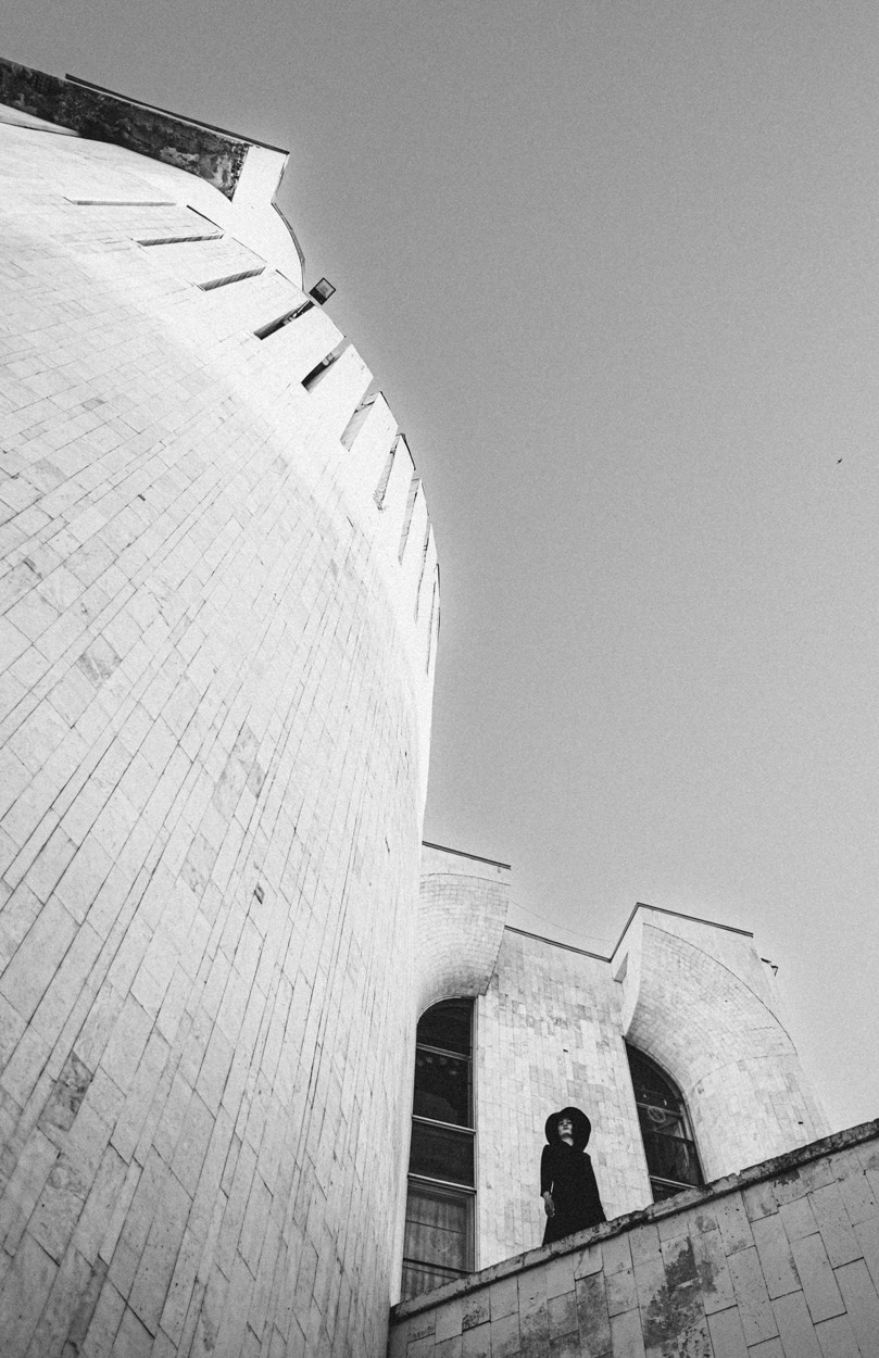 Женщина в чёрной одежде и шляпе стоит у фасада модернистского здания, чёрно-белая фотография с низкой точки