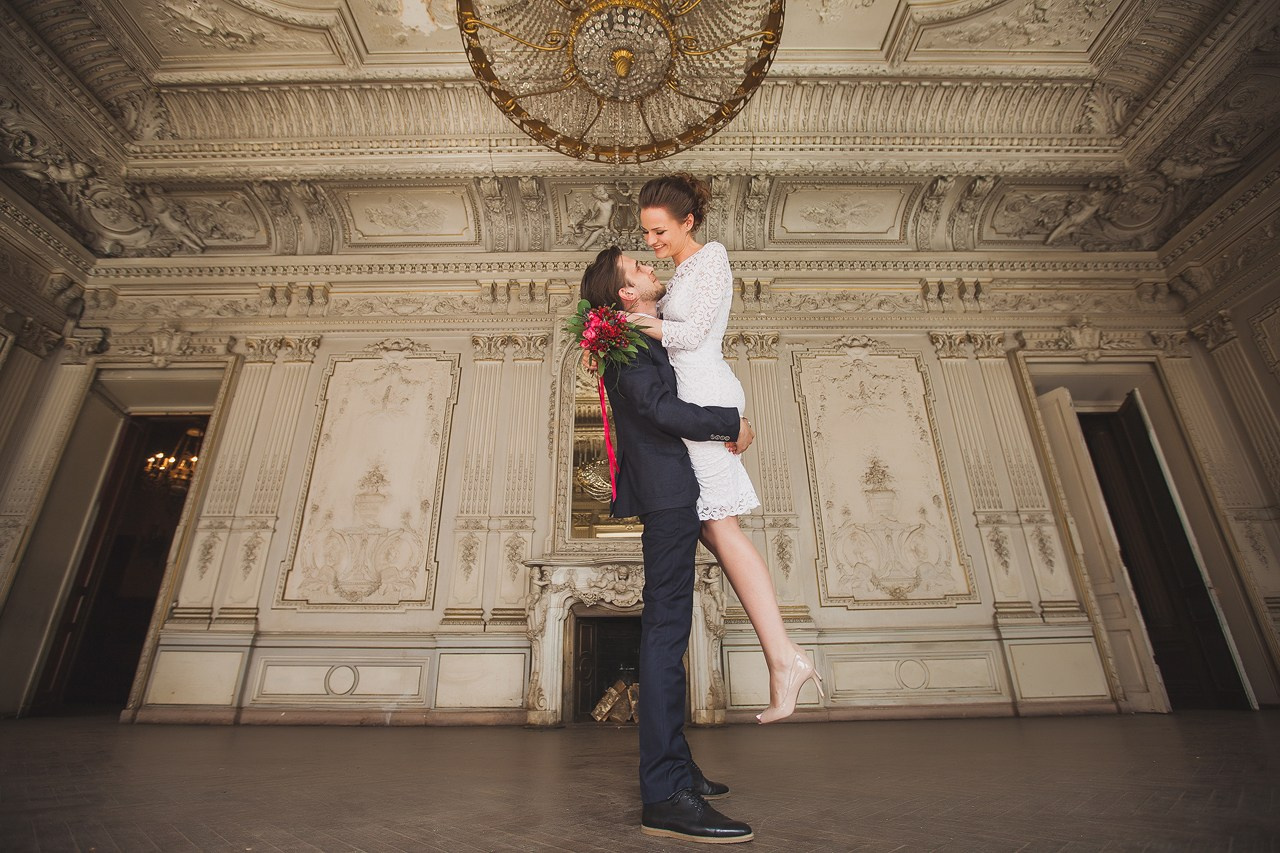 жених приподнимает невесту с красным букетом на фоне большой люстры в старинном зале