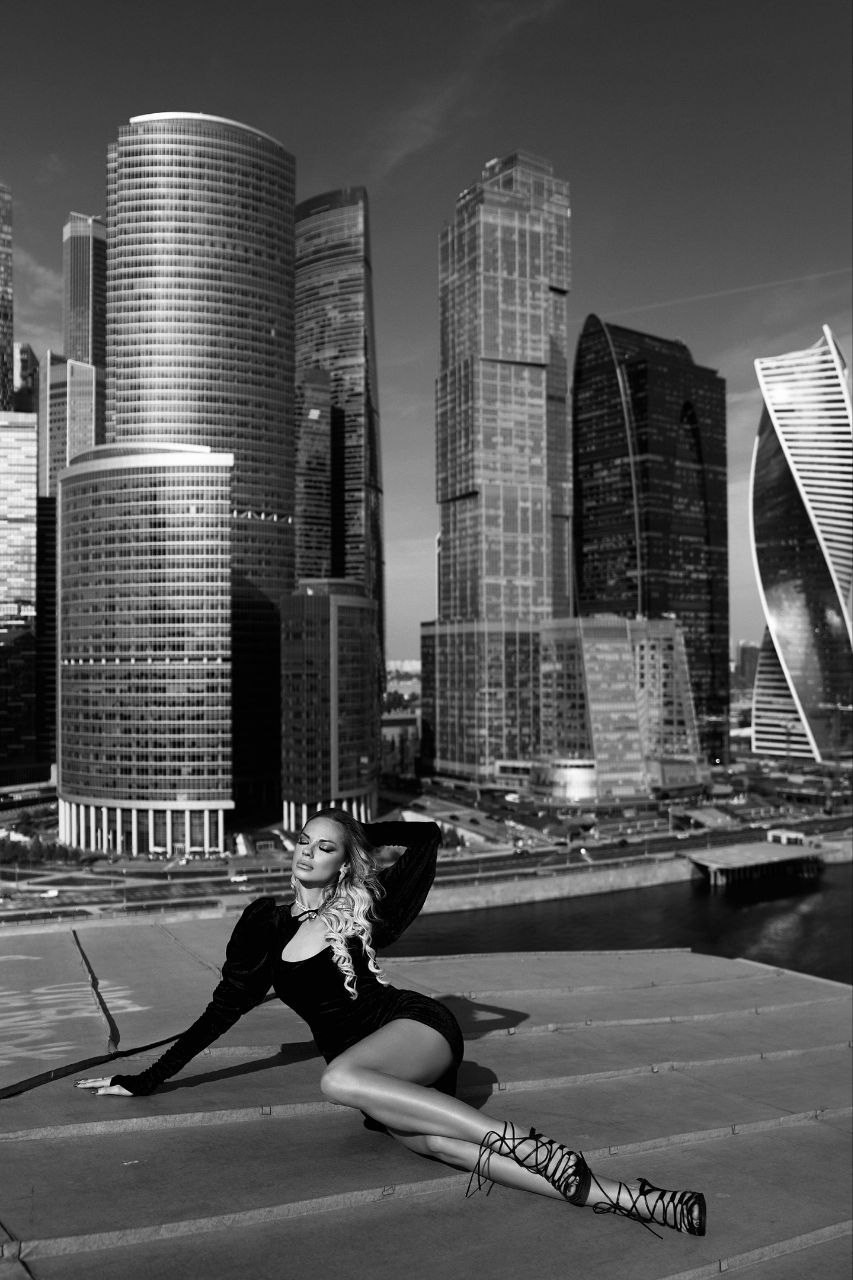 Девушка в черном платье и туфлях на шнуровке лежит на крыше здания на фоне небоскребов