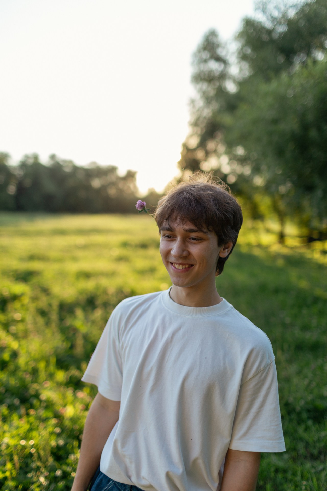 Молодой парень со светлыми волосами и цветком за ухом смеется.