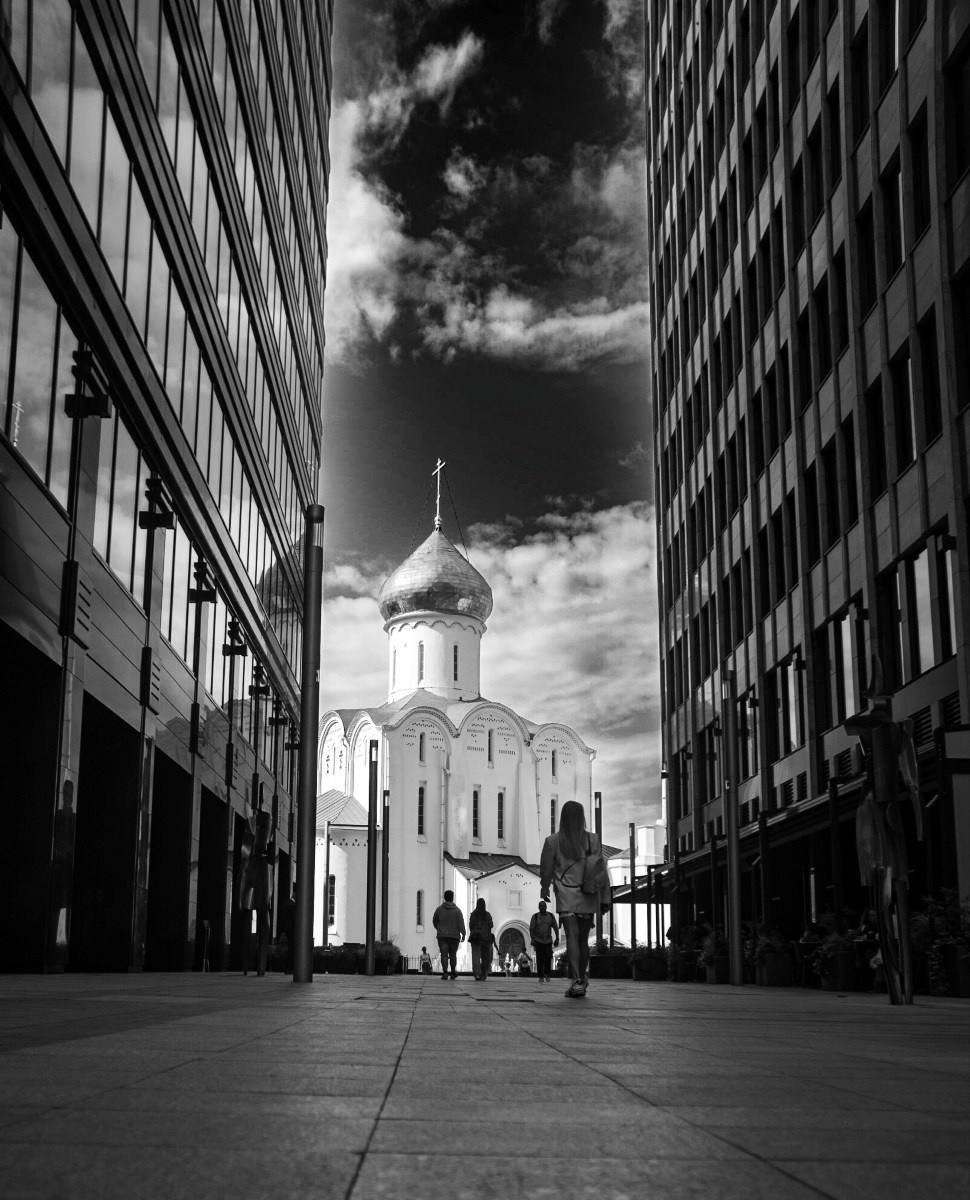 Черно-белая городская фотография: узкая перспектива между двумя современными темными зданиями, в конце аллеи виден белый православный храм с куполом и крестом на фоне драматичного облачного неба, силуэты прохожих идут к церкви, контраст исторической и современной архитектуры Москвы, уличная фотография, Вадим Топорёнок, проект Манифест теней, геометрия города, свет в конце тоннеля, городская археология.