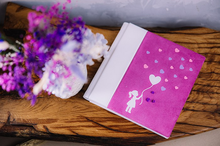 Фотокнига премиум-класса в обложке из ярко-розовой замши и белой кожи. На обложке — силуэт девочки с воздушным шаром в виде сердца. Книга стоит на деревянном спиле рядом с букетом сухоцветов.