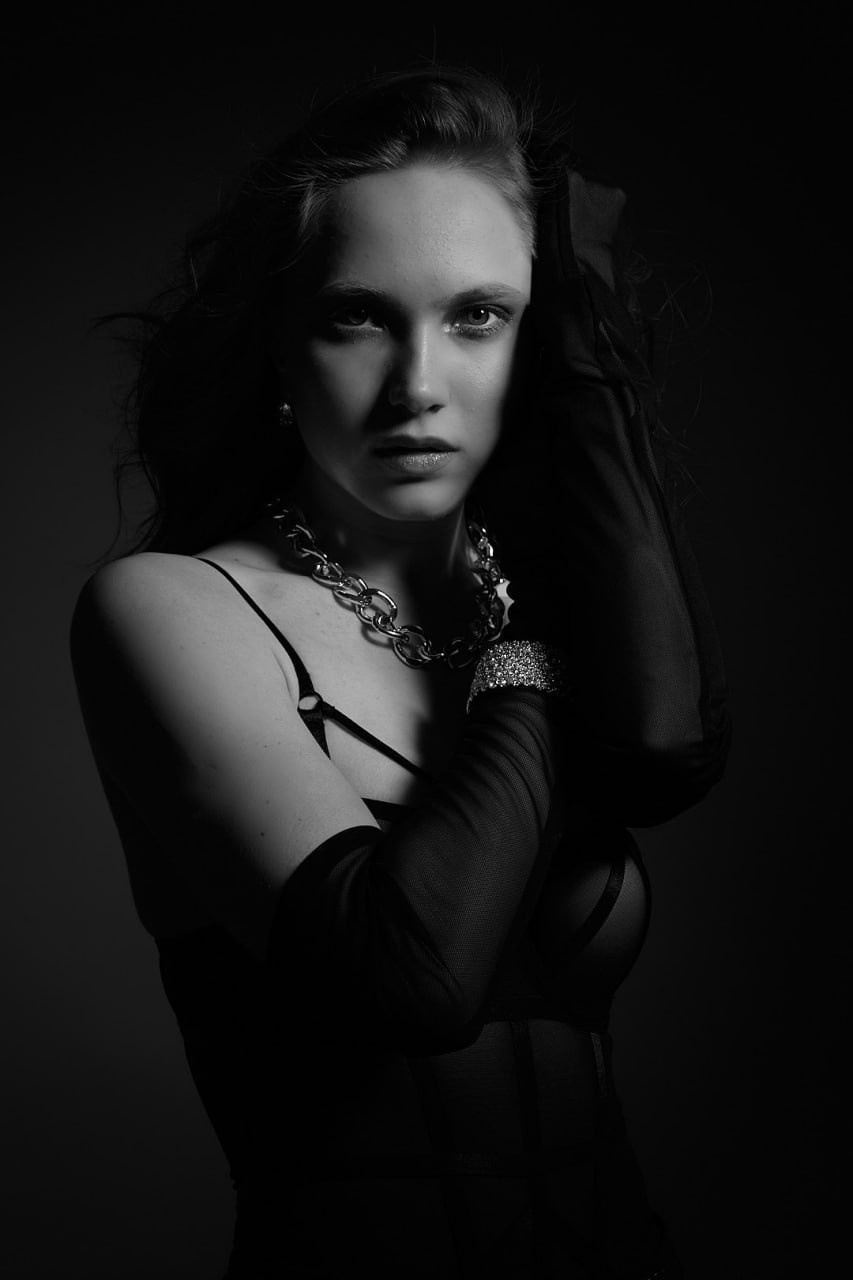 «Чёрно-белый драматичный студийный портрет женщины в полупрозрачном чёрном топе, длинных перчатках, на шее — массивная серебряная цепочка и блестящий браслет. Резкое освещение падает на её лицо, создавая контрастный образ, вдохновлённый фильмами в стиле нуар».