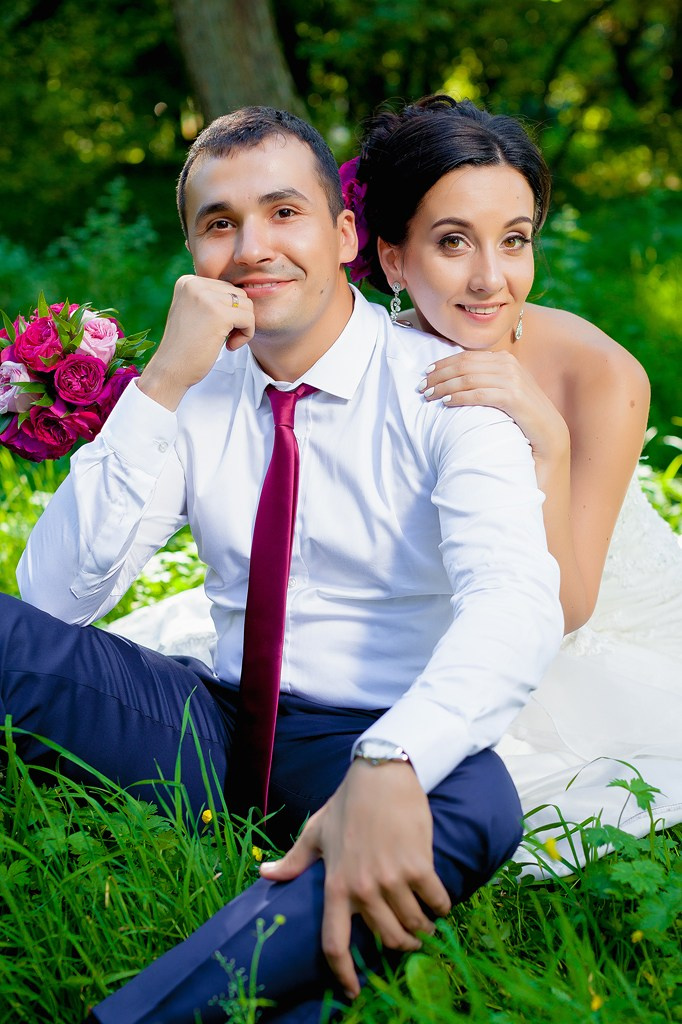 жених и невеста с малиновым букетом на зеленой траве при ярком солнечном свете