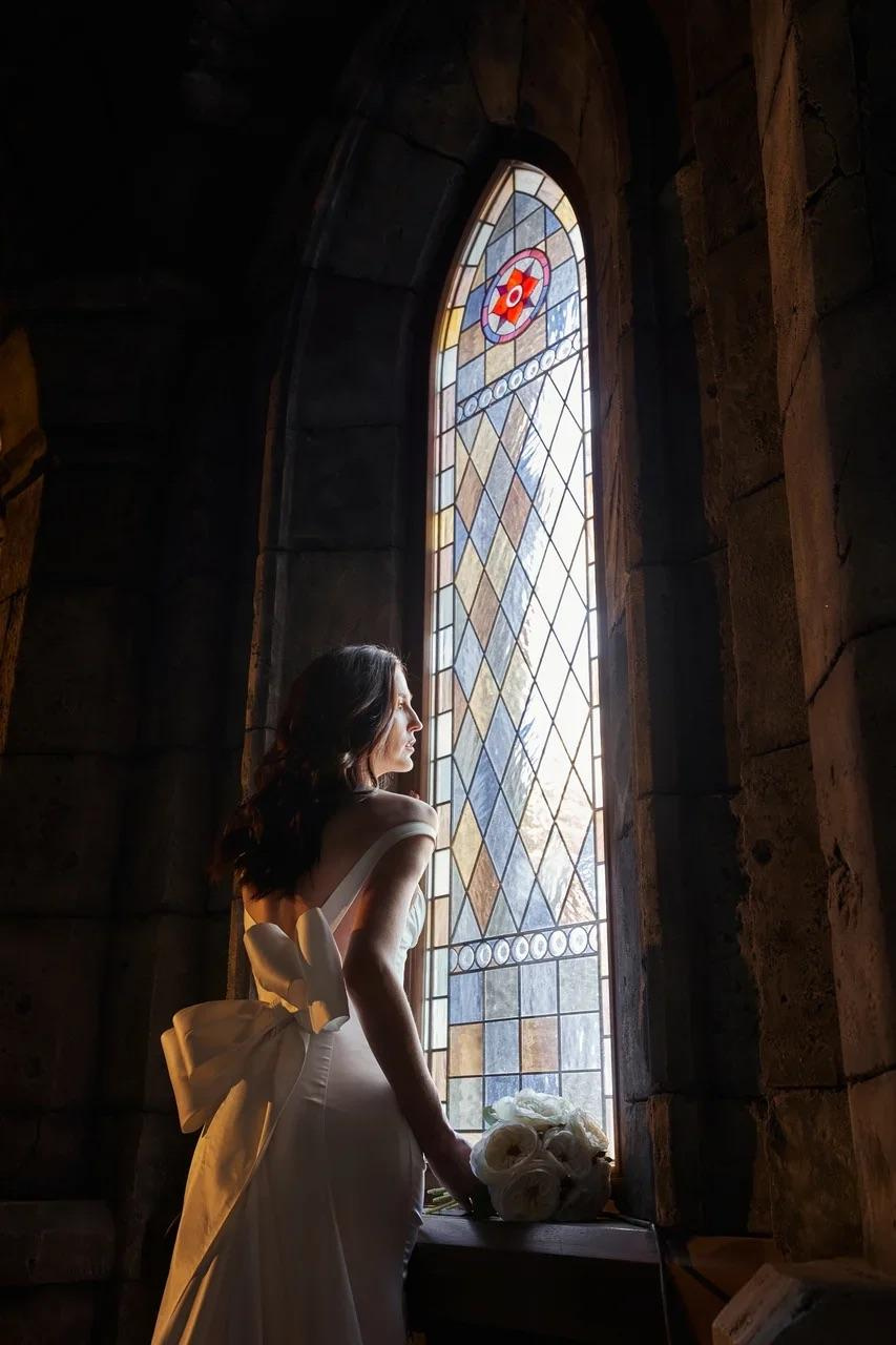 Невеста у большого витражного окна внутри замка Гарибальди, где мягкий свет подчеркивает торжественный характер съёмки.