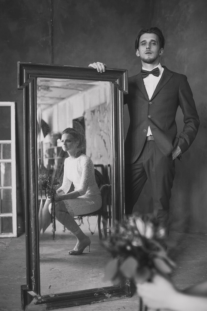 жених в темном костюме и бабочке держит большое зеркало с отражением невесты
