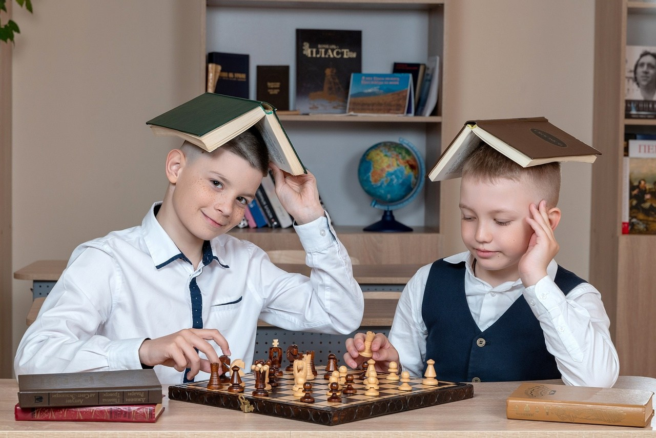 Двое ребят сидят за партой с учебниками на голове позируя фотографу.