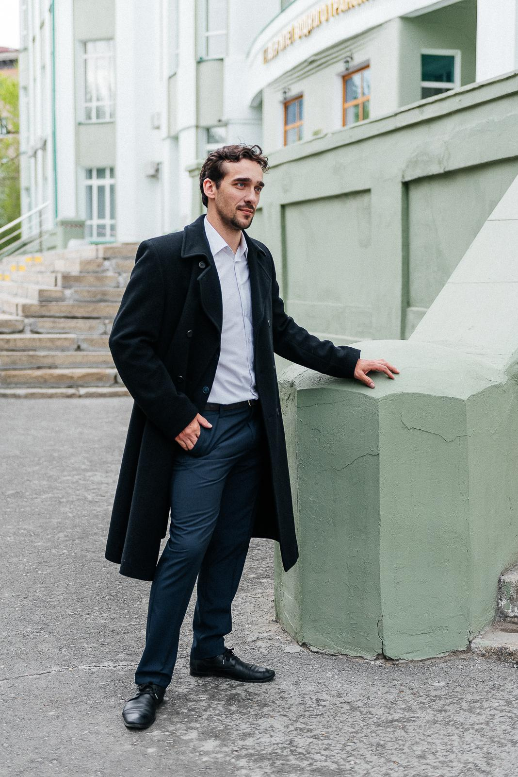 парень в черном пальто стоит у здания с советской архитектурой, фотосессия в городе,  фотограф новосибирск