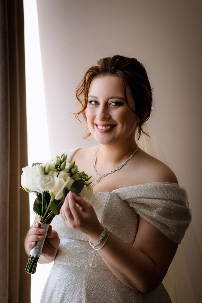 Невеста у окна смотрит в кадр и улыбается