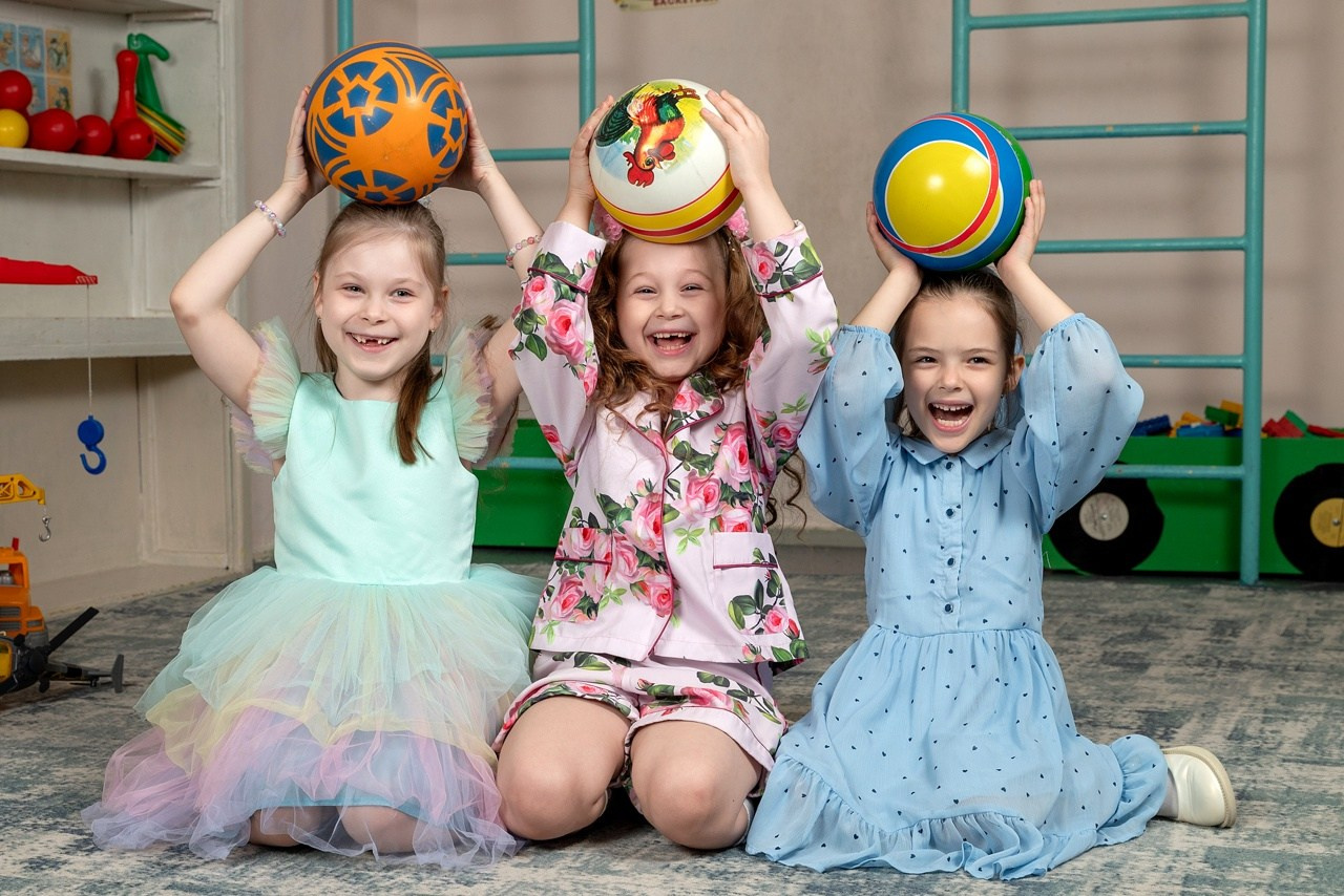 Девочки позируют с разноцветными мячиками на головах улыбаясь фотографу.