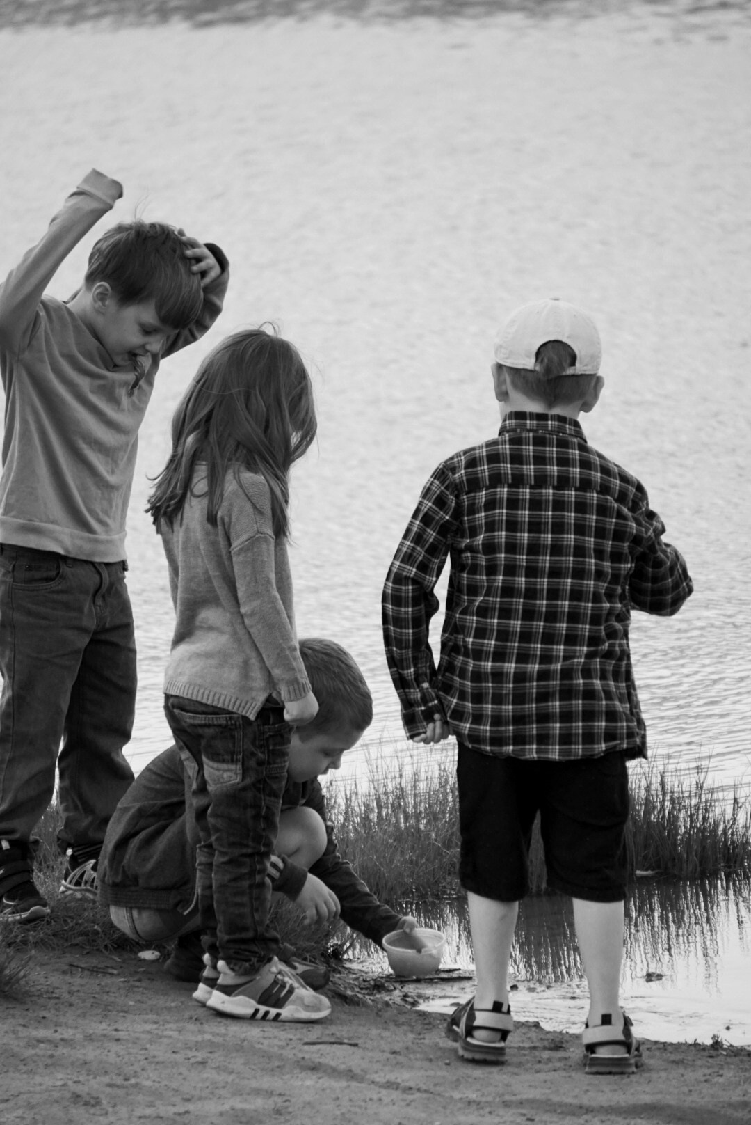 Четверо детей играют у водоёма: один мальчик зачерпывает воду миской, остальные наблюдают; чёрно-белая фотография.