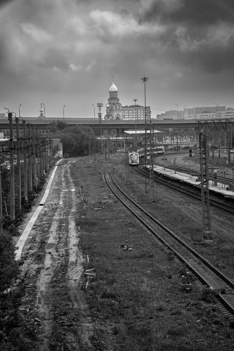 Черно-белая городская фотография: изогнутые железнодорожные пути уходят вдаль через мокрую насыпь с лужами, слева забор и контактная сеть, на заднем плане сквозь облака виден купол храма, драматичное небо, уличная фотография Москвы, Вадим Топорёнок, Манифест теней, археология душ, геометрия пути, урбанистический пейзаж.