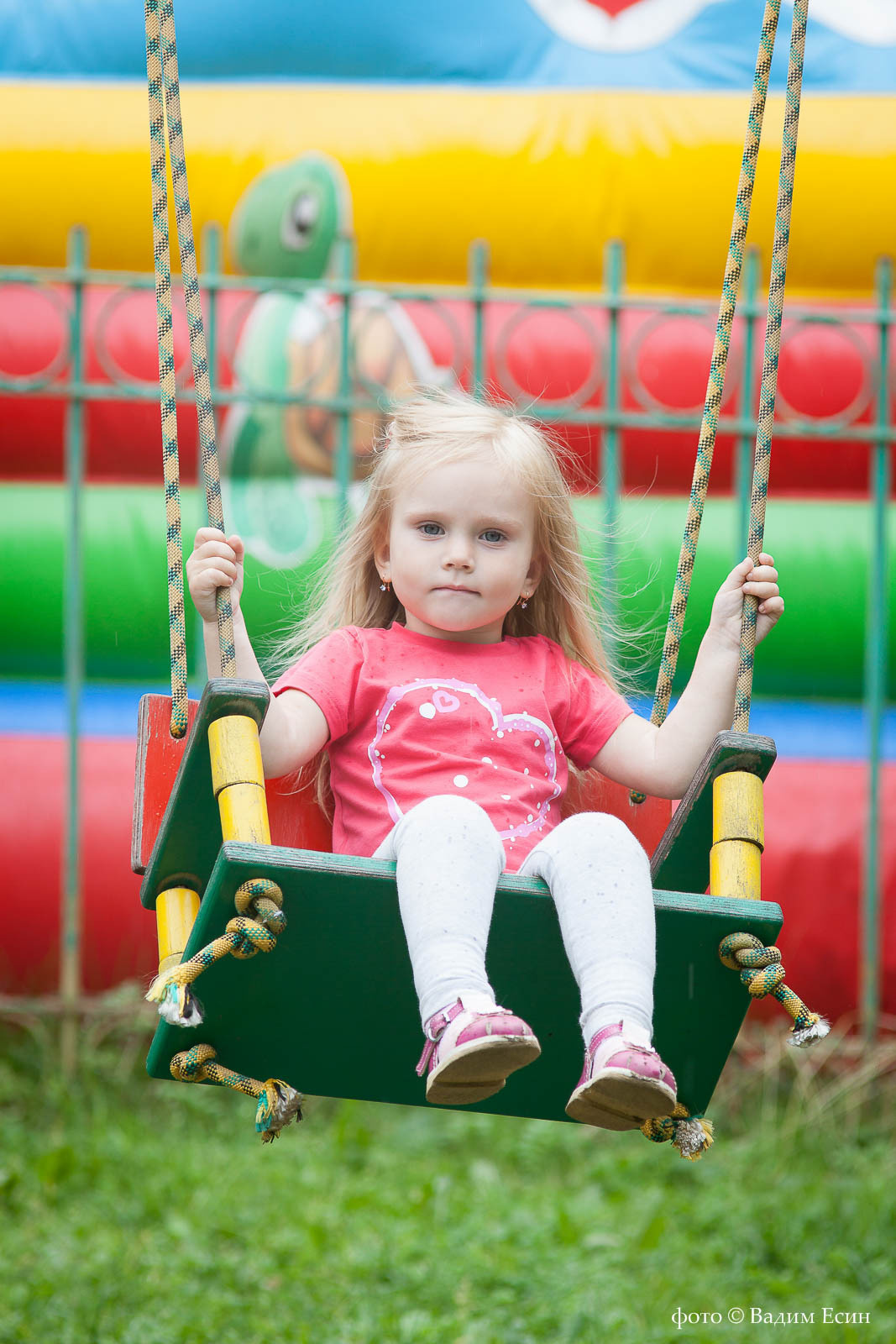Девочка в красной футболке качается на качелях . Сзади разноцветный надувной батут.