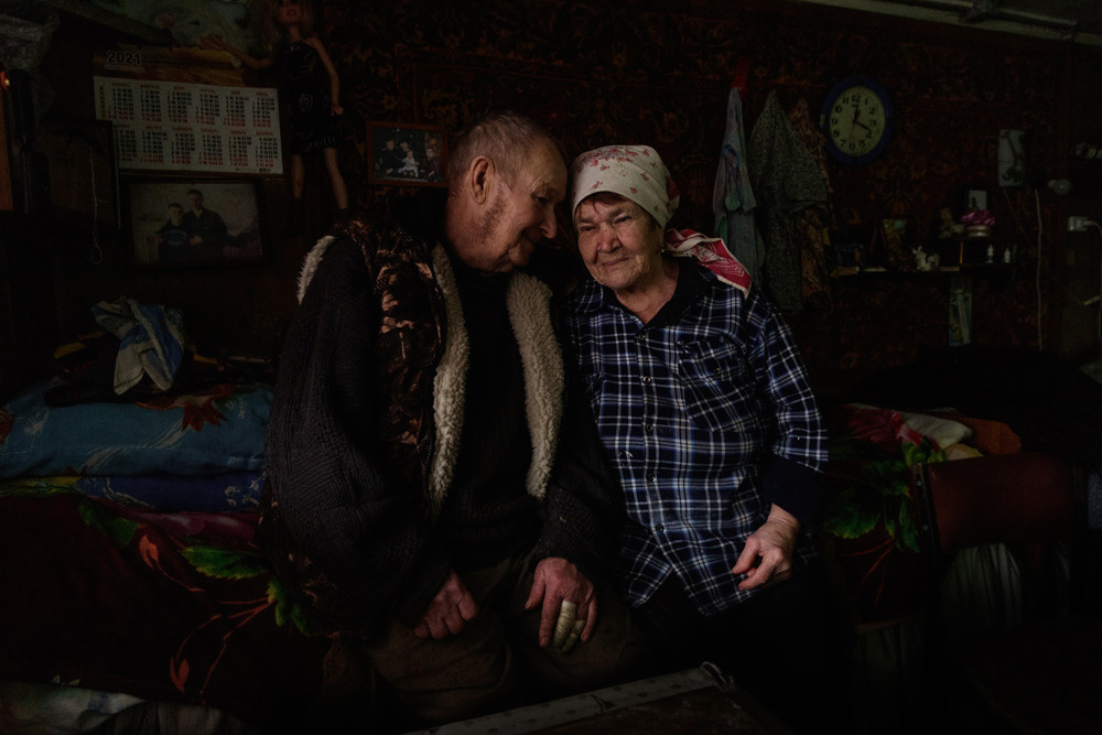 Два пожилых человека мужчина и женщина сидят на кровати в тёмной комнате.