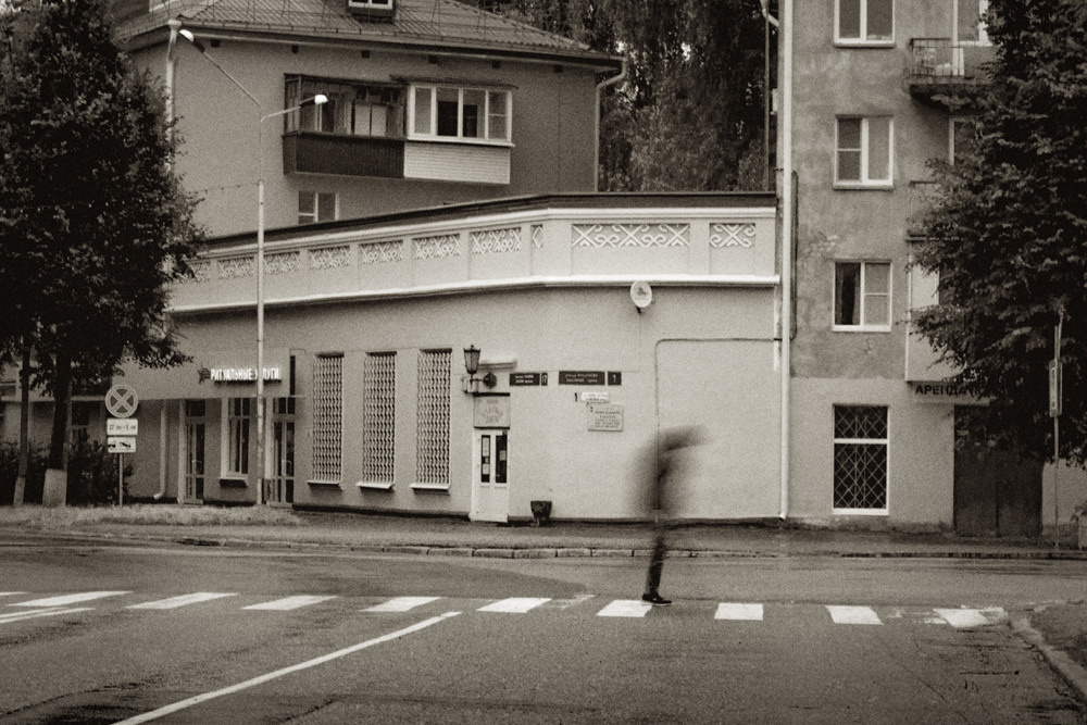 Размытая фигура человека на пешеходном переходе на фоне салона ритуальных услуг.