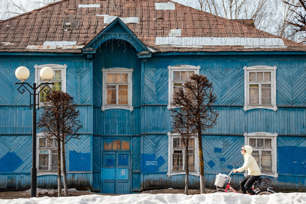 Глоубой старый деревянный дом и проезжающая на велосипеде девушка с ведром.