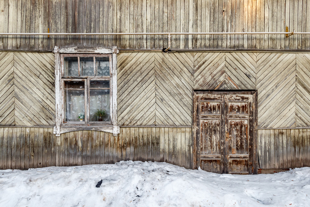 Фасад старого дома. Подъезд в сугробе, а в окне цветы и ветка пихты.