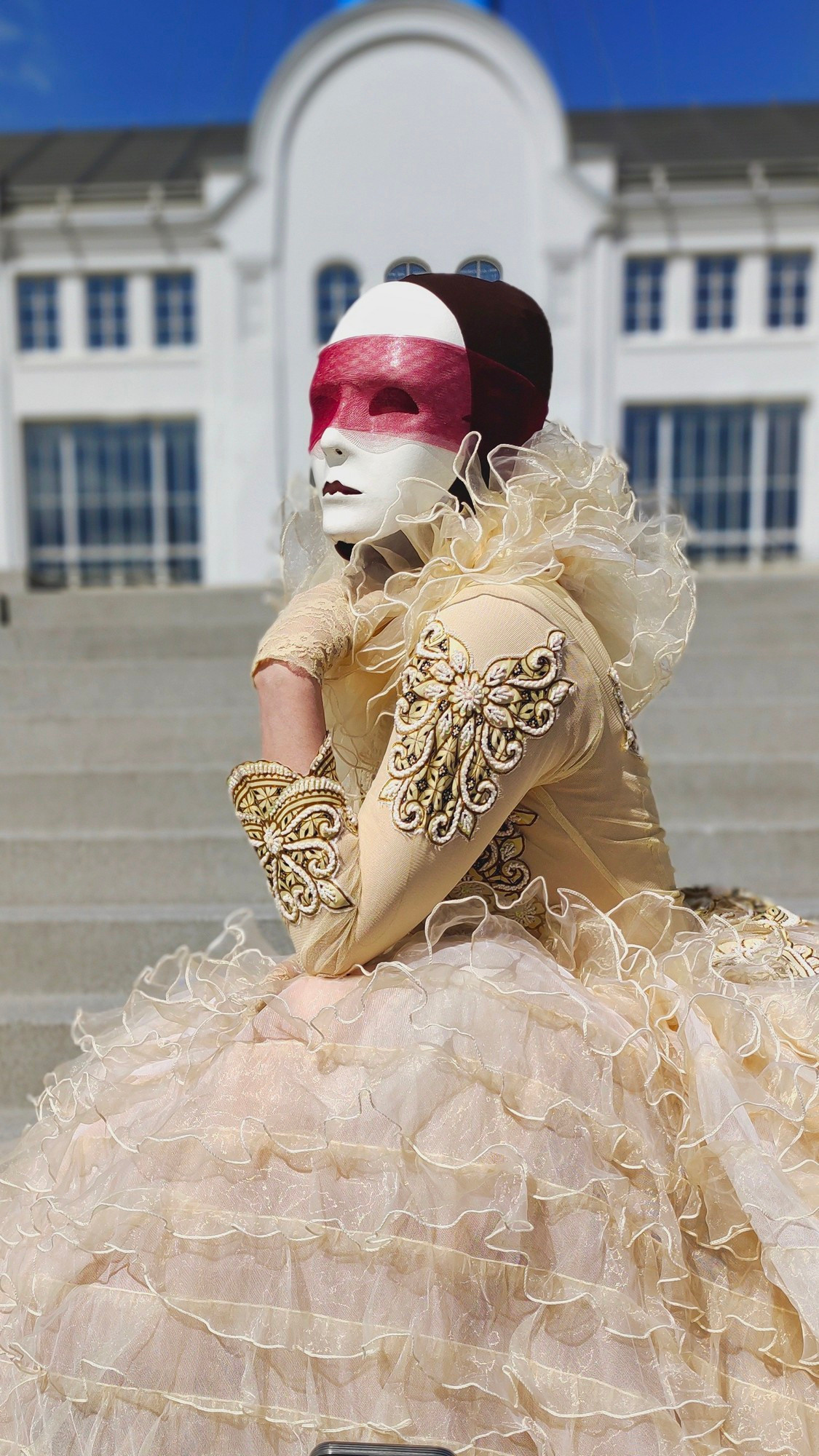 На лестнице на фоне здания сидит артист мим в светлом платье и белой маске.