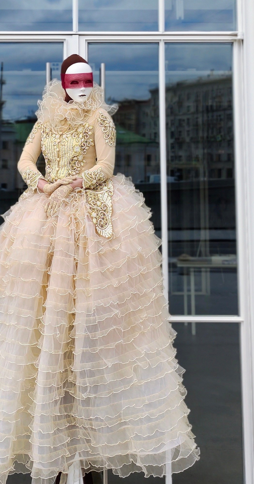 Светлая Пешка на ходулях стоит у окна большого белого здания.