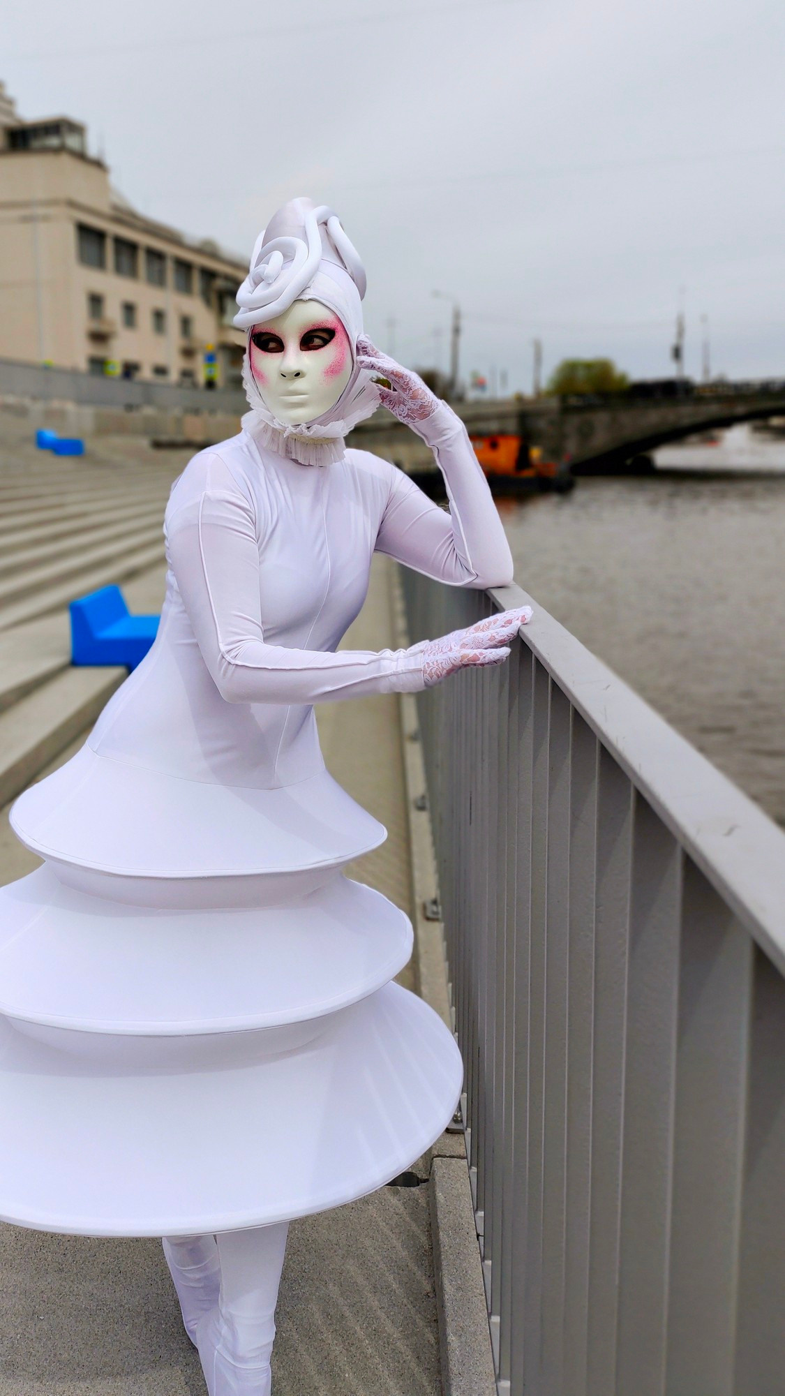 Мим в белом образе и белой маске опирается на ограждение на набережной реки Москва.