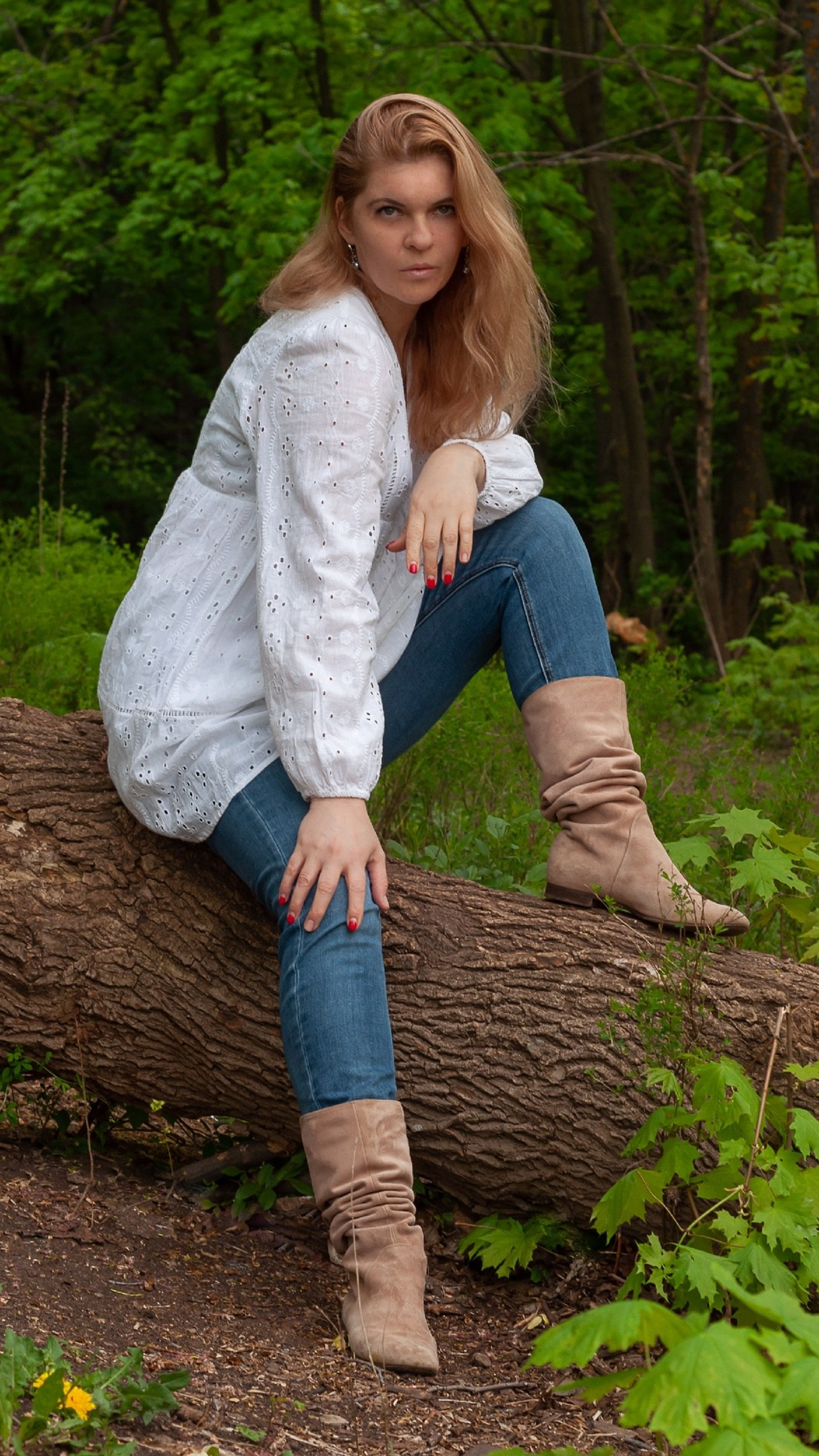 Женщина сидит на упавшем дереве в лесу в белой блузе и светлых сапогах — спокойствие и природная простота.