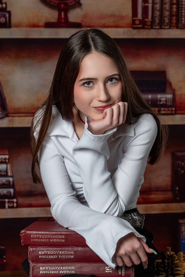 Девочка в белой блузке сидит в классе и красиво смотрит в объектив фотоаппарата.