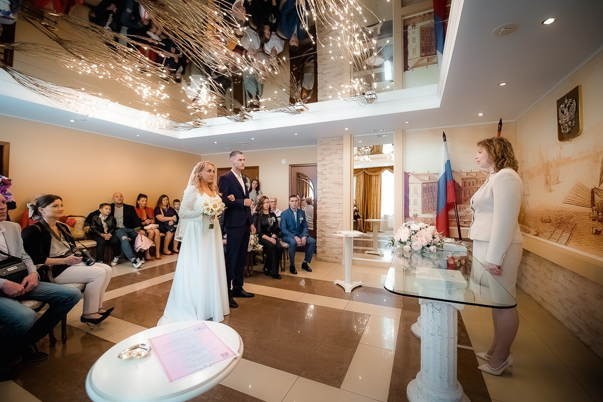 Жених и невеста стоят и смотрят на регистратора во время регистрации брака в ЗАГСе на Васильевском острове в Санкт-Петербурге. Горизонтальный ростовой кадр.