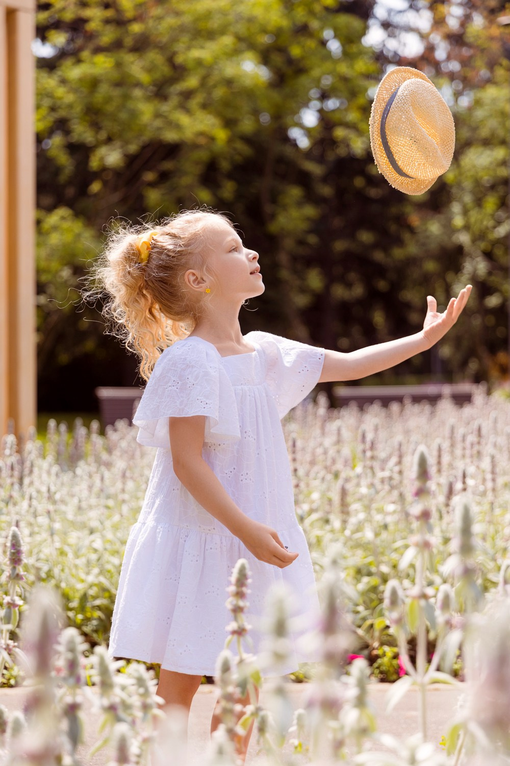 Девочка стоит в клумбе цветов, подбрасывая вверх шляпку.