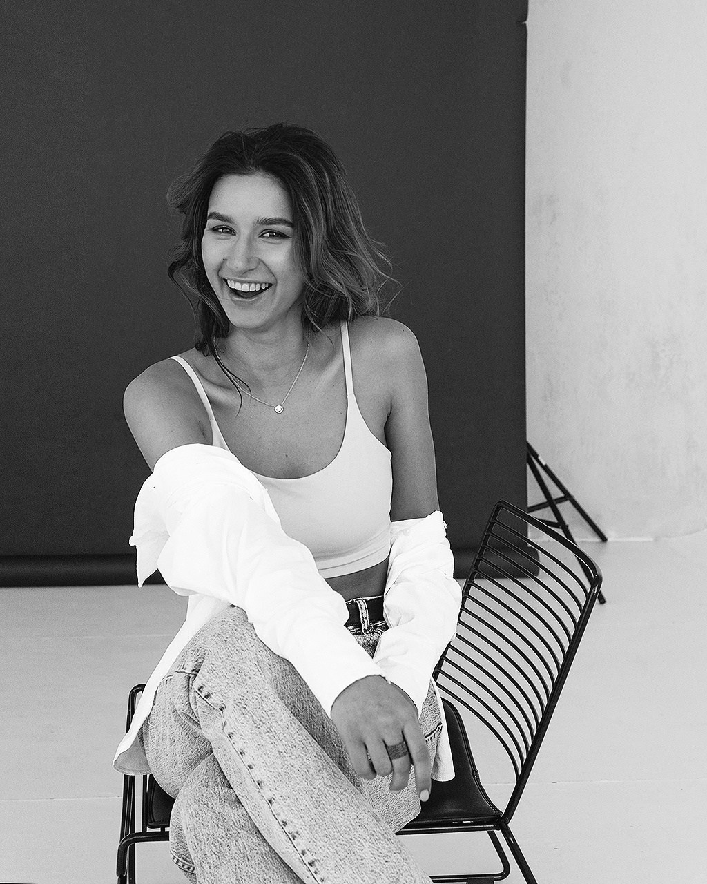 Черно белый портрет девушки на сером фоне, которая улыбается, сложила нога на ногу. Девушка в топике, белой рубашке и светлых джинсах.