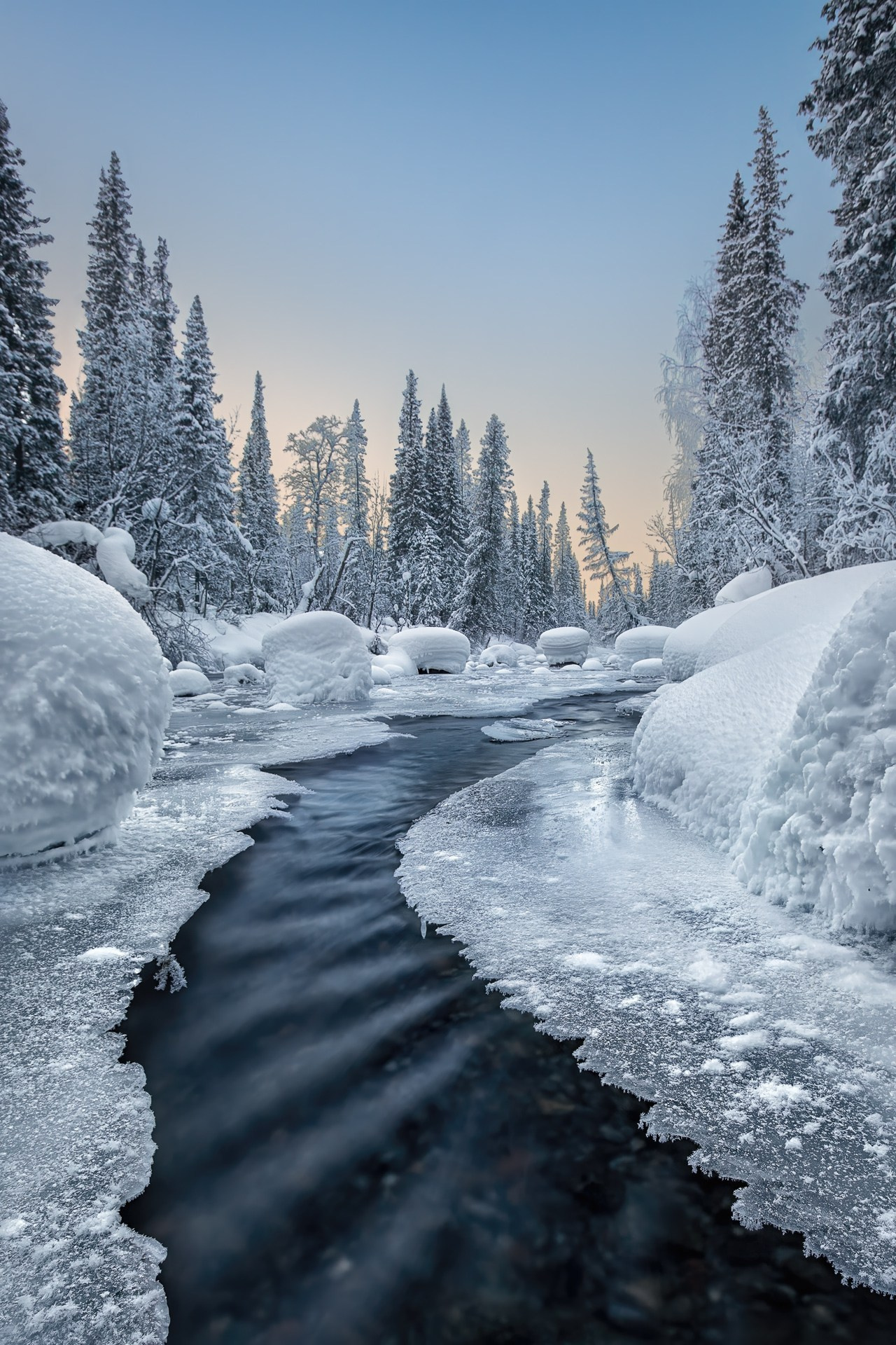 Речка с ледяными берегами уходит вдаль, на горизонте высокие заснеженные ели