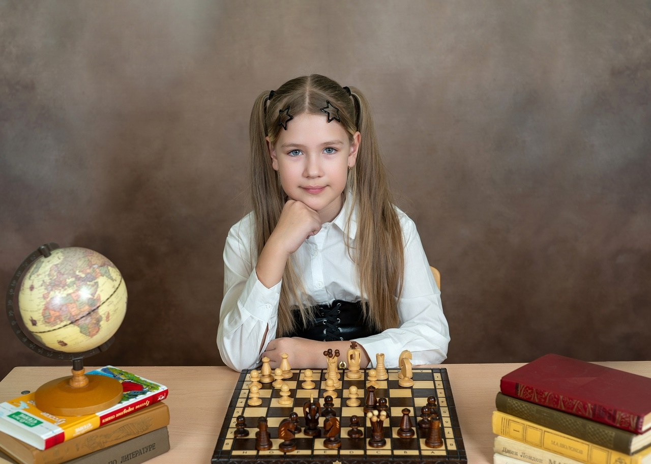 Девочка фотографируется сидя за партой, с глобусом и шахматами.