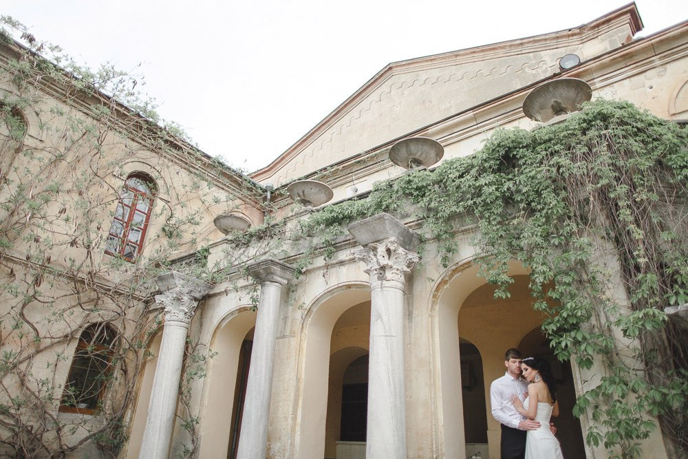 Молодожены идут по аллее, перекрытой свадебным аркой, украшенной цветочными гирляндами