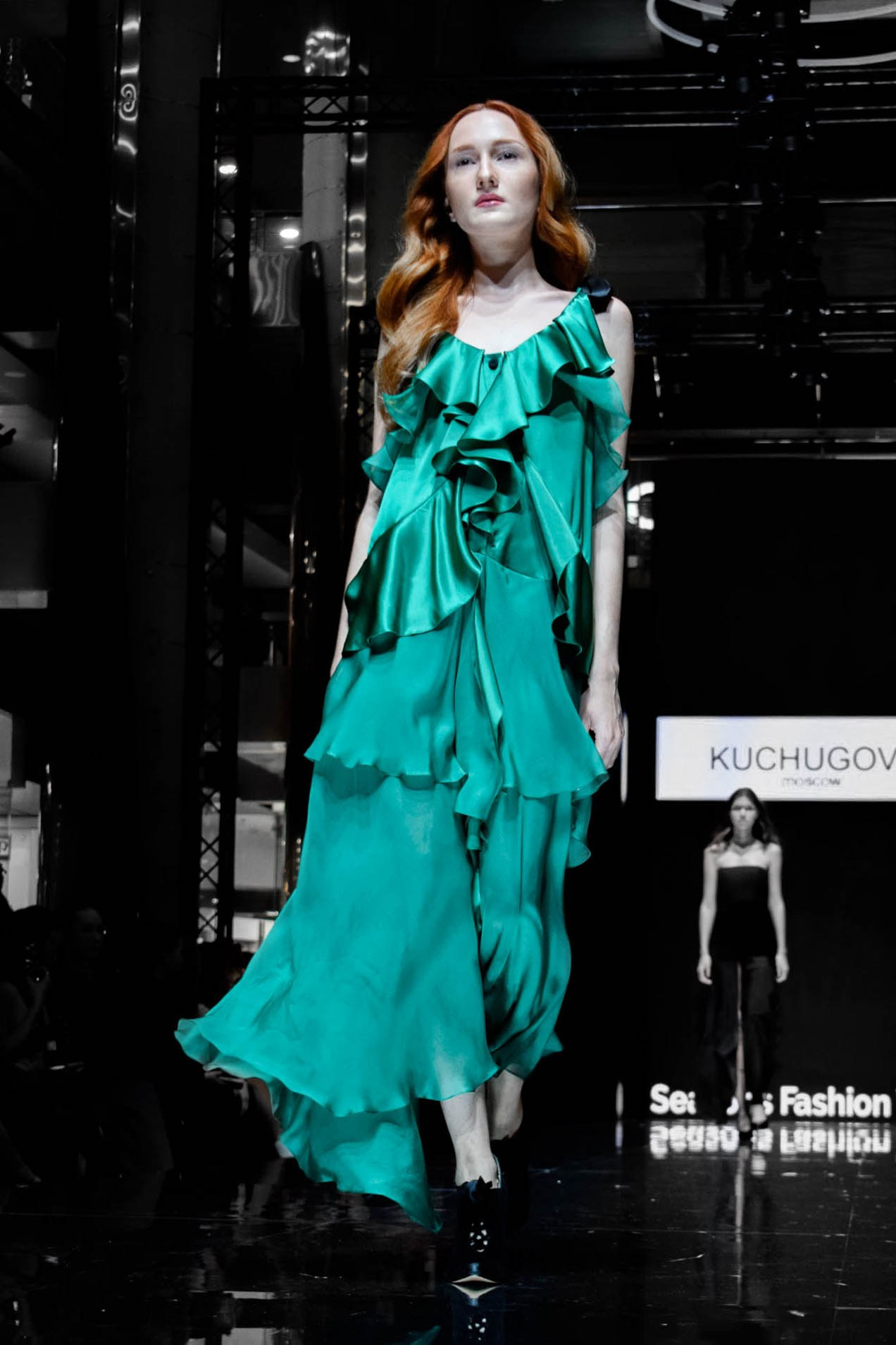 Рыжая девушка в зеленом платье идет по темному подиуму 