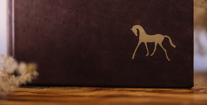 Крупный план золотого тиснения на темно-коричневой коже: изящный силуэт лошади, подчеркивающий премиальный статус индивидуального фотоальбома.