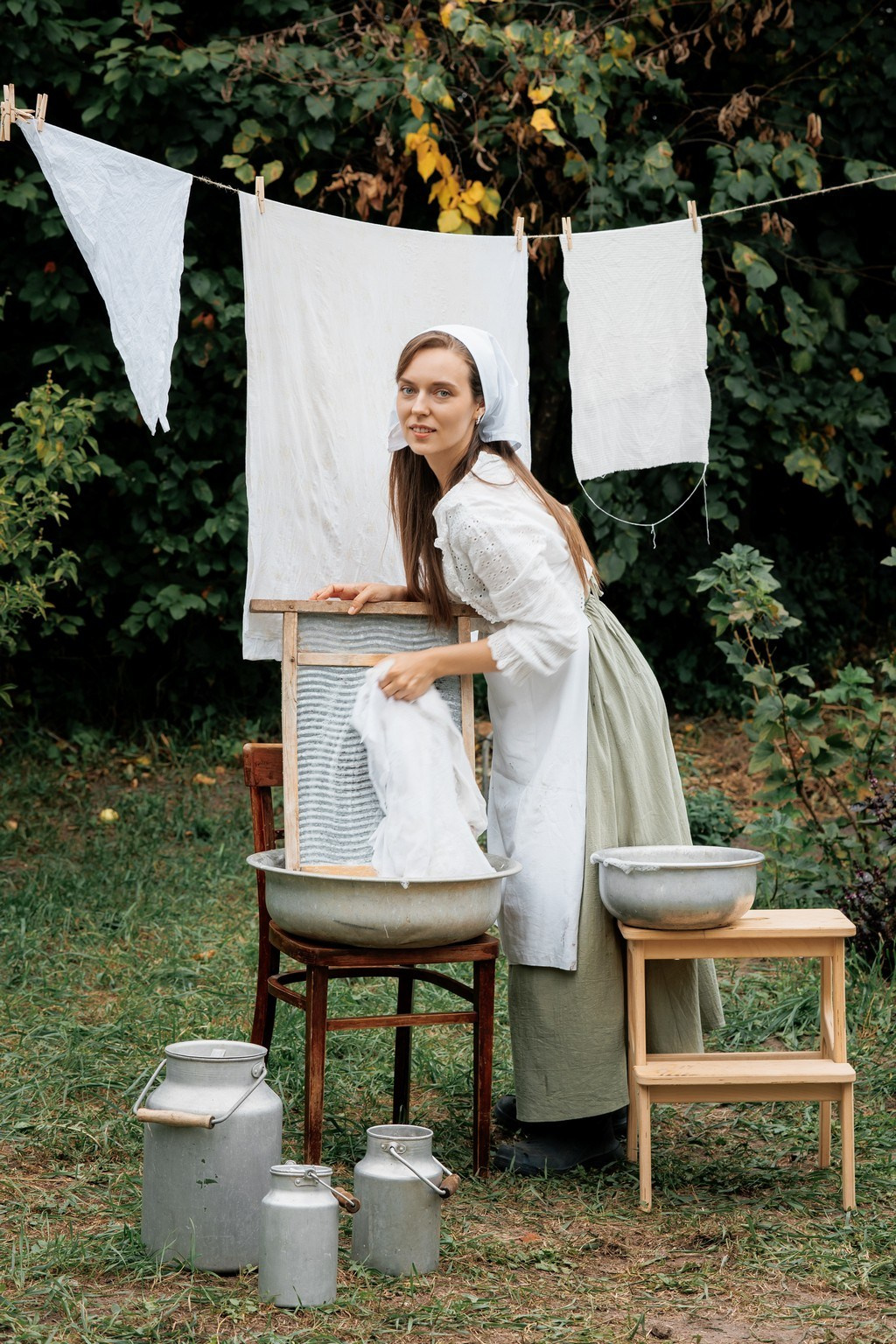Женщина стирает белое бельё вручную в металлическом тазу с помощью деревянной стиральной доски в саду.