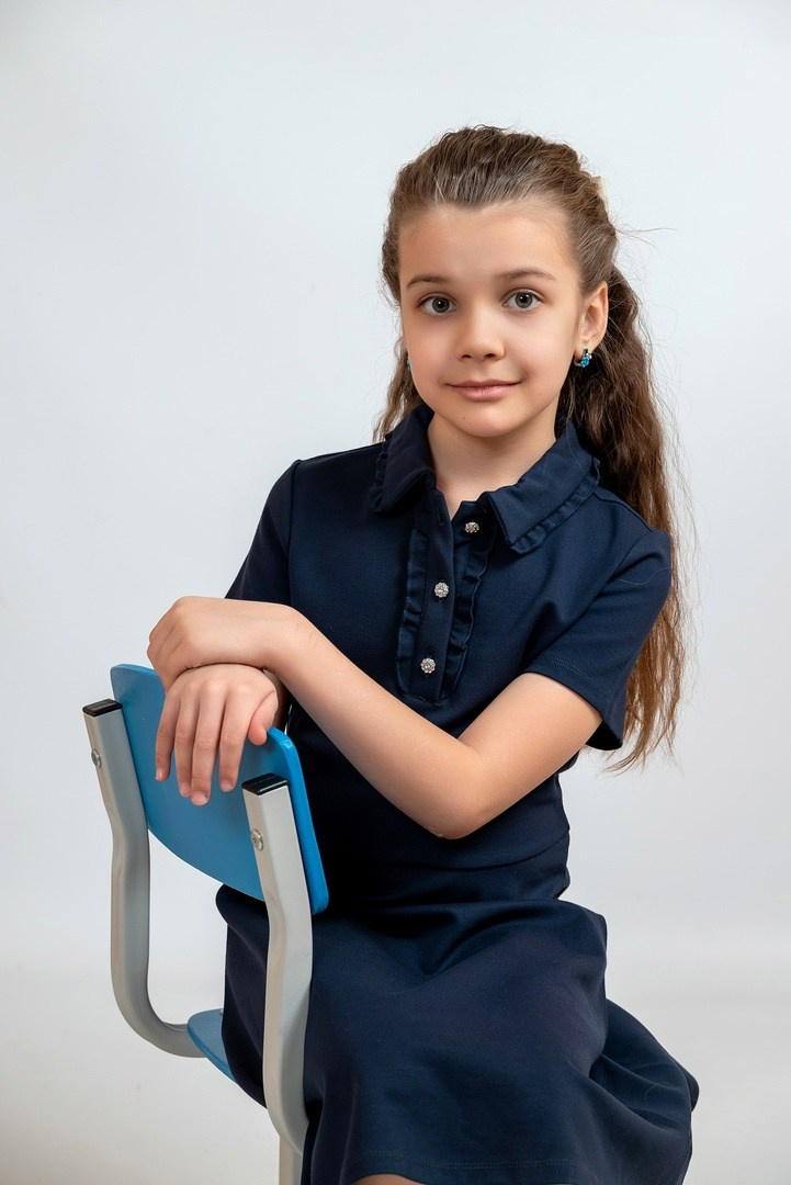 Симпатичная девочка в черном платьице, фотографируется сидя на стульчике.