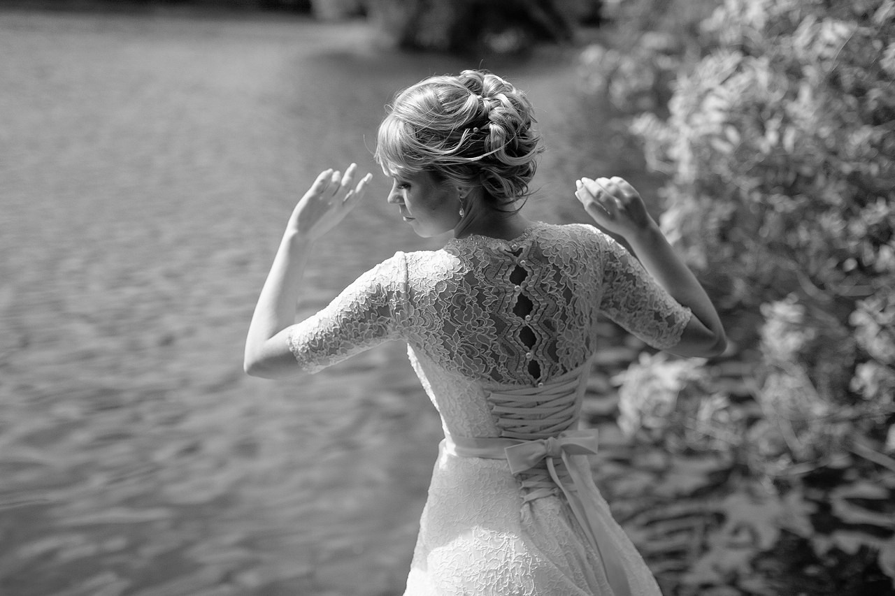 невеста в дизайнерском платье с кружевной спиной позирует на фоне воды 