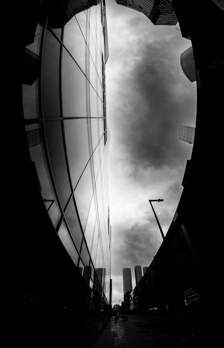 Черно-белая уличная фотография с эффектом круглой виньетки: вид сквозь тёмный проём на стеклянный фасад здания, отражающий грозовое небо с тяжёлыми облаками, внизу мокрый асфальт и силуэты высоток, портальная композиция, городская архитектура Москвы, Вадим Топорёнок, проект Манифест теней, геометрия города, урбанистическая фотография, свет и тень в городе.