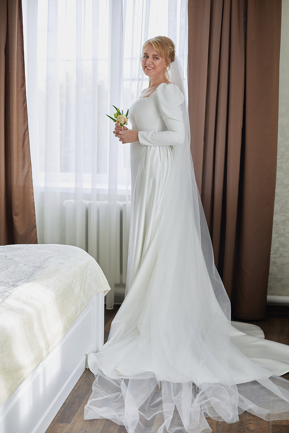 Утро невесты. Невеста с бутоньеркой в руках стоит у окна. Невеста улыбается. Фотограф. Тимашевск. Кубань. Краснодарский край.